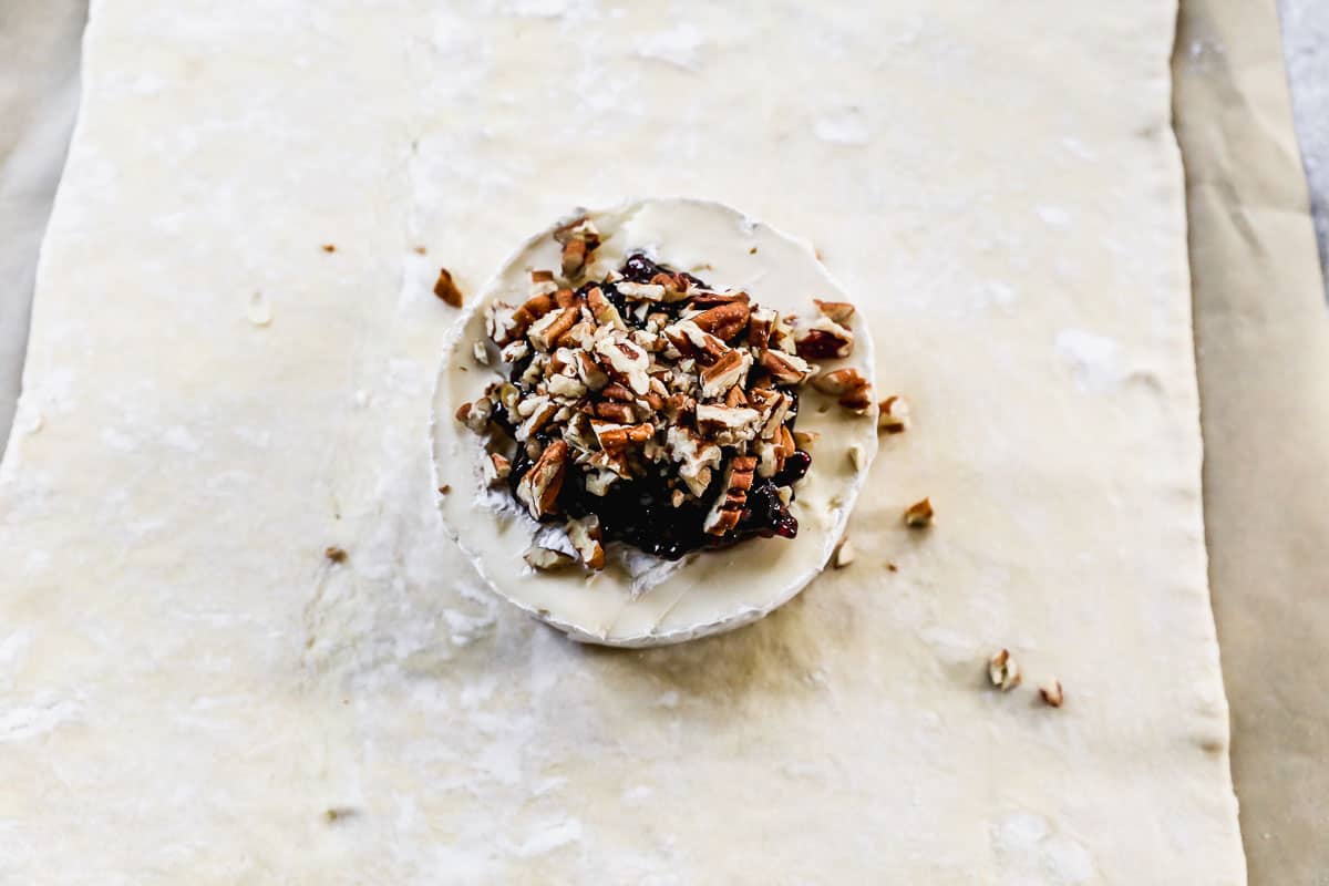 A wheel of brie with jam and pecans on top of it, almost ready to be wrapped in puff pastry.