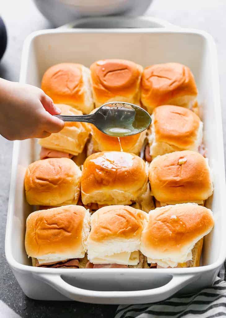 Butter dijon sauce being spooned over ham and cheese sliders in a baking dish.