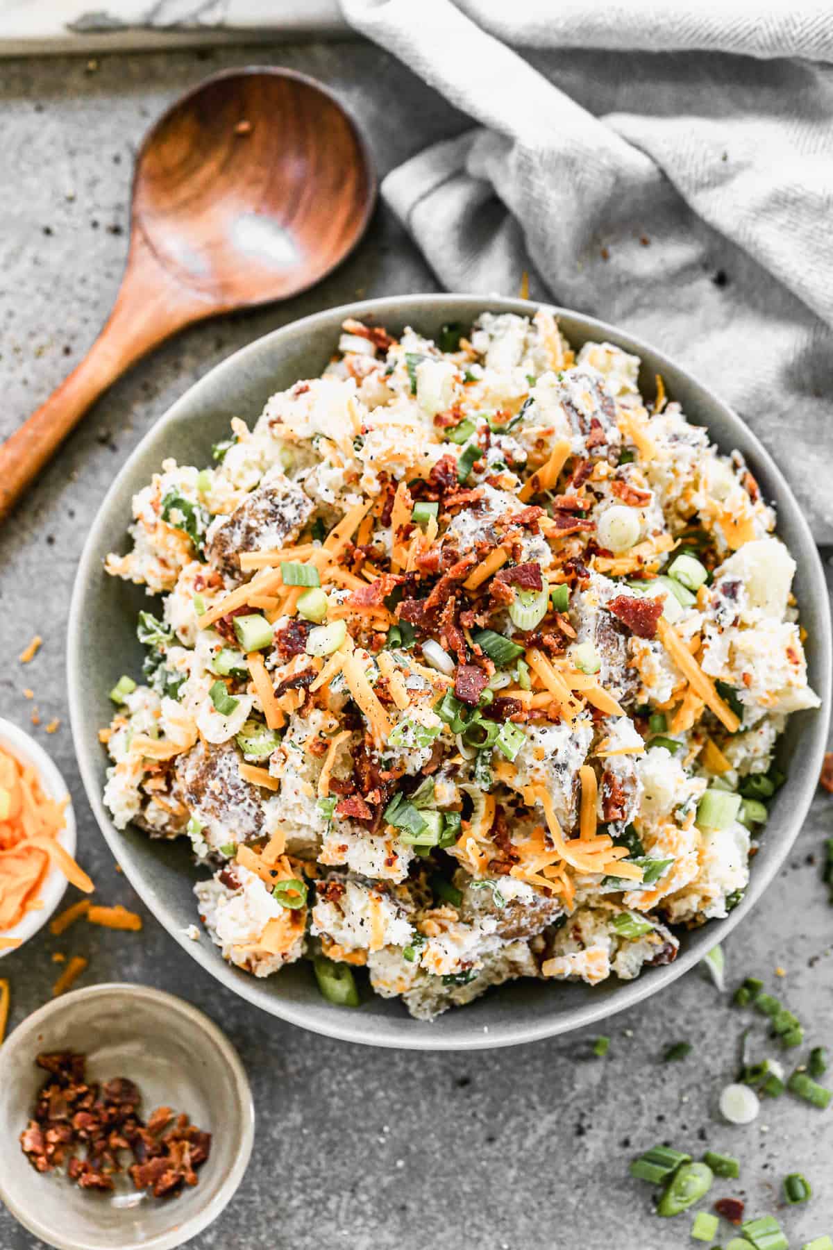 A serving bowl filled with Baked Potato Salad, topped with cheddar cheese, crumbled bacon, and sliced green onions.