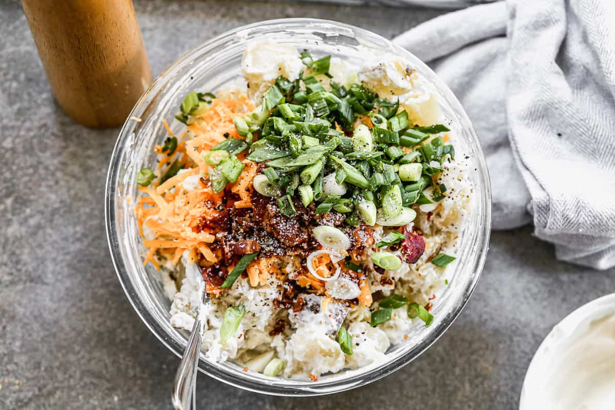 Chopped baked potatoes tossed in a sour cream sauce and topped with cheddar cheese, bacon, and green onion for a loaded potato salad recipe.