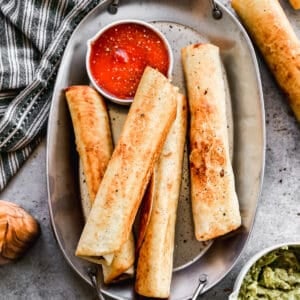 A platter of four bean and cheese burritos on a platter with a side of salsa and a bowl of guacamole.