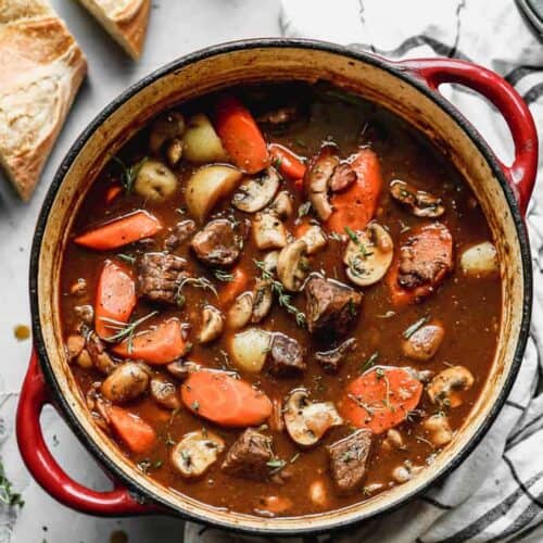 A large pot of beef stew with carrots, potatoes and mushrooms and sliced baguette on the side.