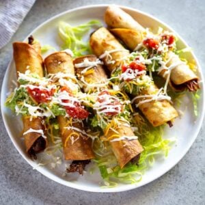 Five beef taquitos fanned out on a plate topped with guacamole, sour cream and salsa.