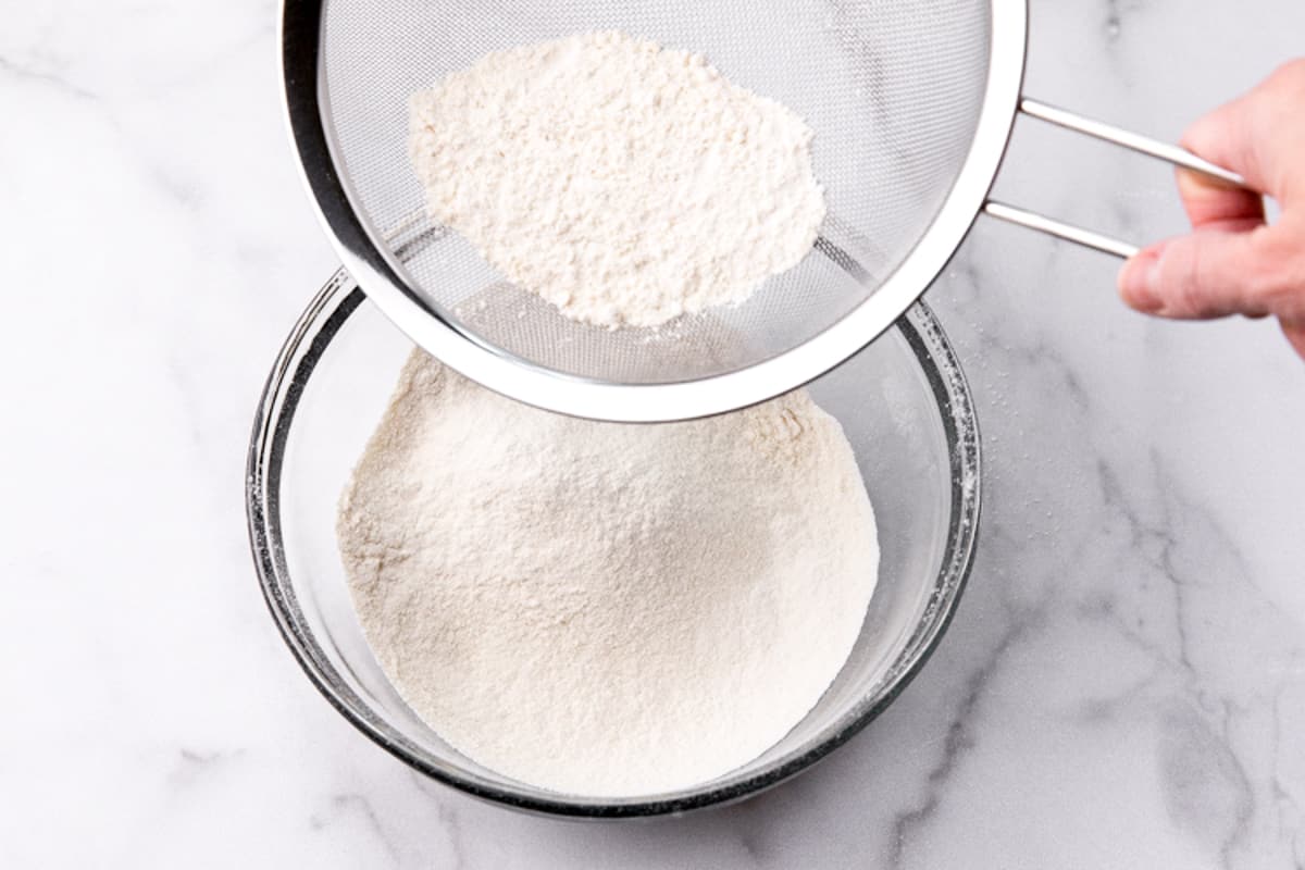 A mesh wire strainer sifting the dry ingredients into a glass bowl for Belgian Waffles.