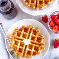 Belgian waffles on plates with butter, syrup, and powdered sugar next to a bowl of strawberries.