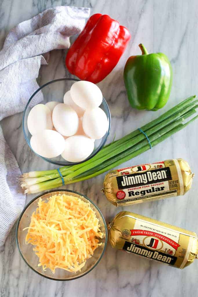 An overhead photo of the ingredients for breakfast casserole including eggs, red and green bell pepper, green onion, sausage and shredded cheddar cheese.