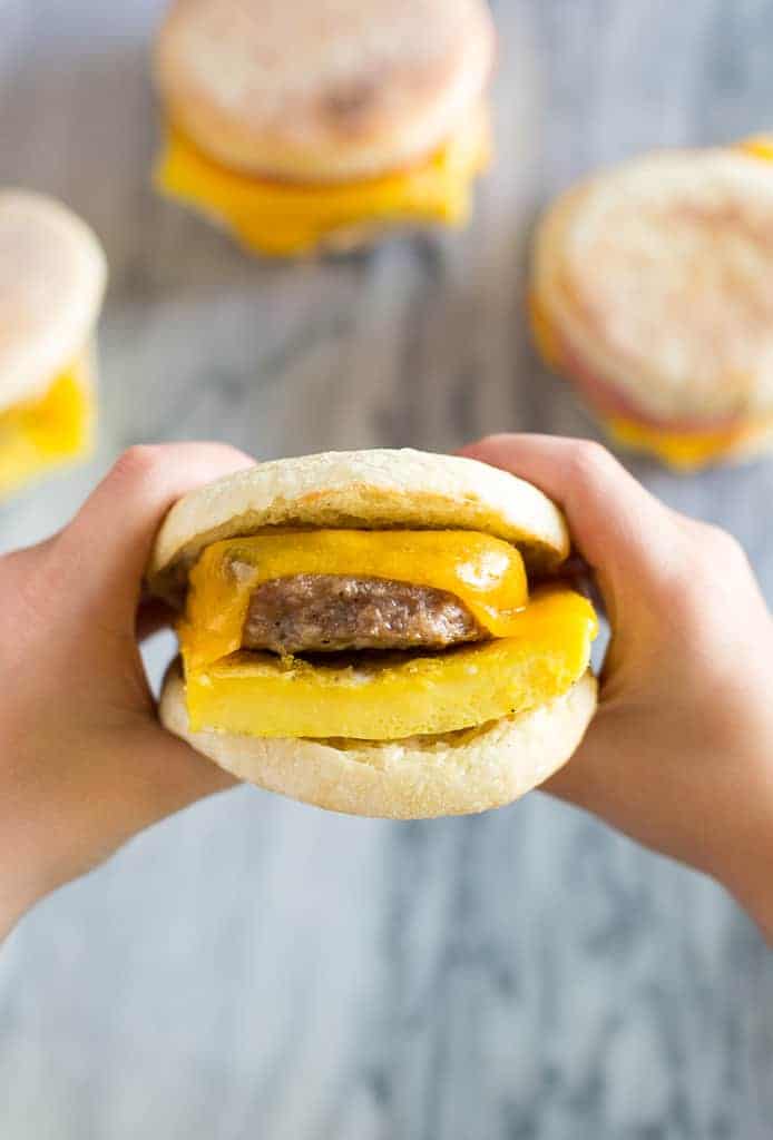 Overhead photo of hands holding an English muffin breakfast sandwich with sausage, egg and cheese and more sandwiches in the background.