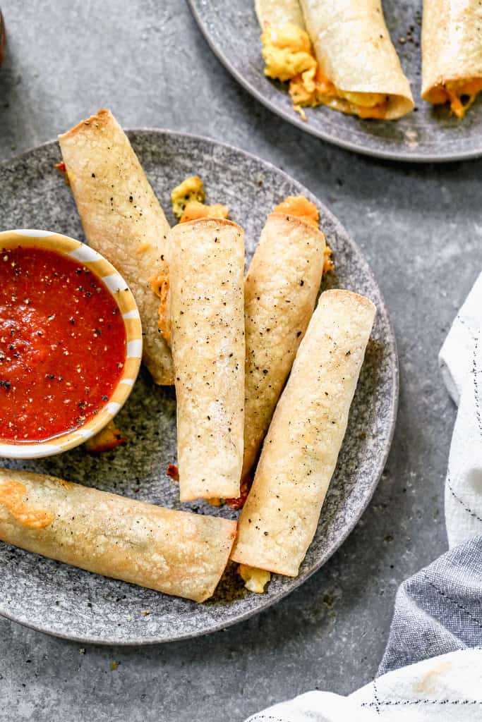 Five breakfast taquitos on a plate with a small bowl of salsa.