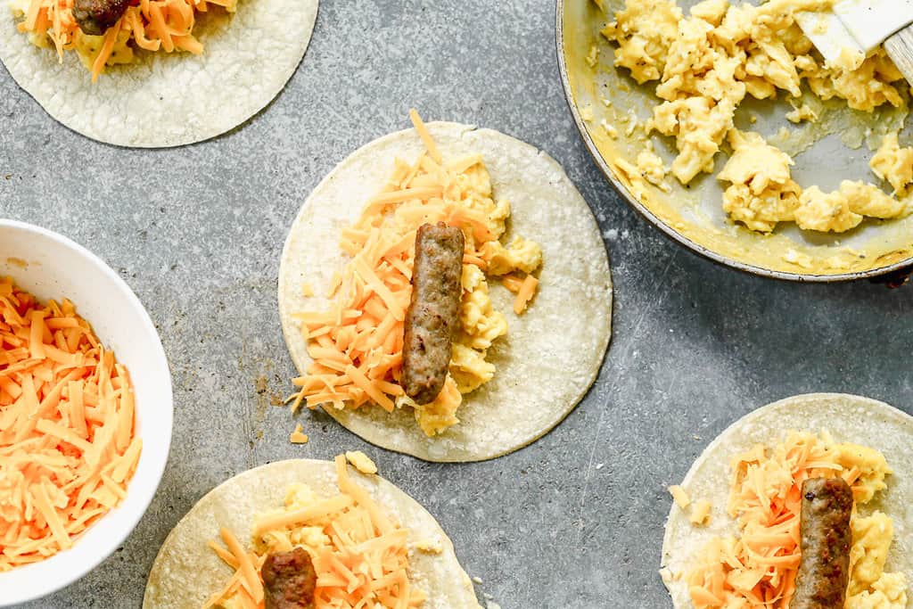 Easy breakfast taquitos being assembled with scrambled eggs, cheese, and sausage on a corn tortilla.