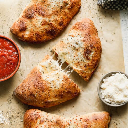 Three homemade Calzones on a piece of parchment paper, with the middle one cut in half showing the cheese pulling apart.