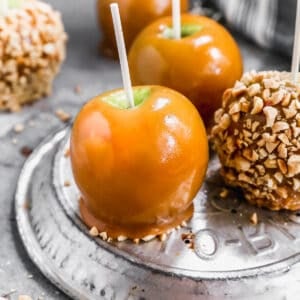 A close up image of three caramel apples on an upside down pie plate, one of them covered with chopped nuts.