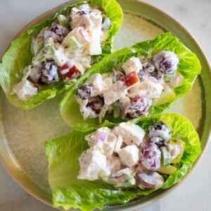 Three lettuce cups on a plate filled with chicken salad mixture with cut grapes and apples in it.