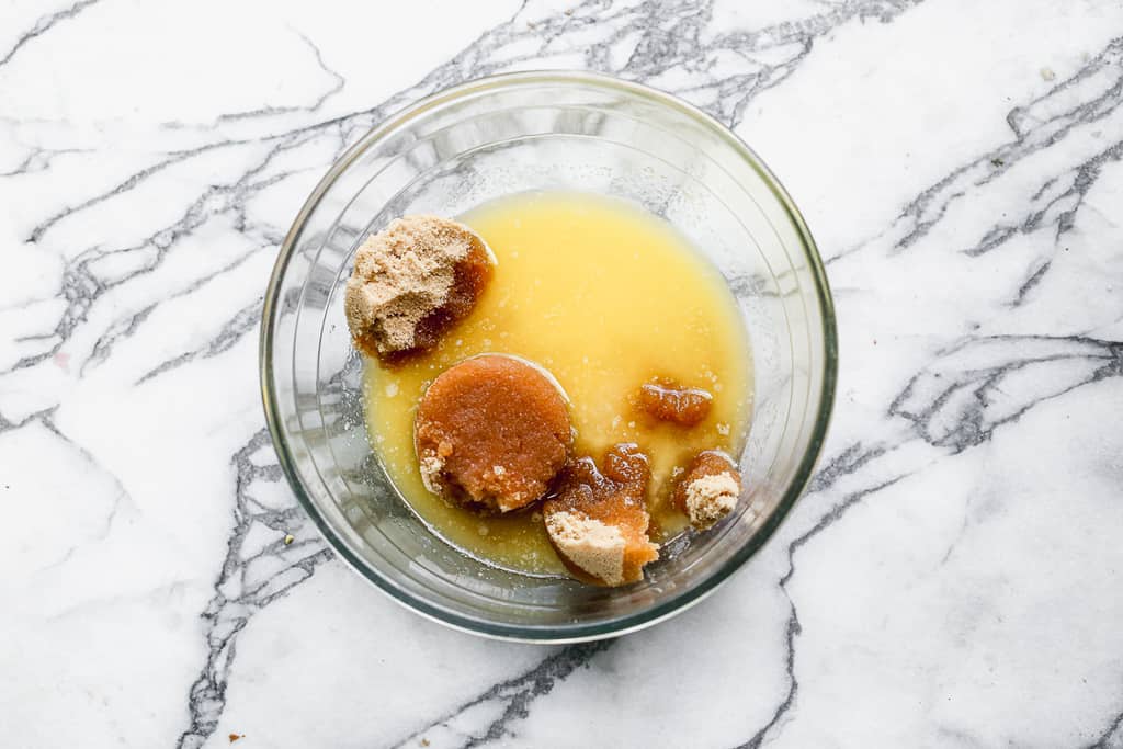 A clear glass mixing bowl with melted butter, granulated sugar and brown sugar.