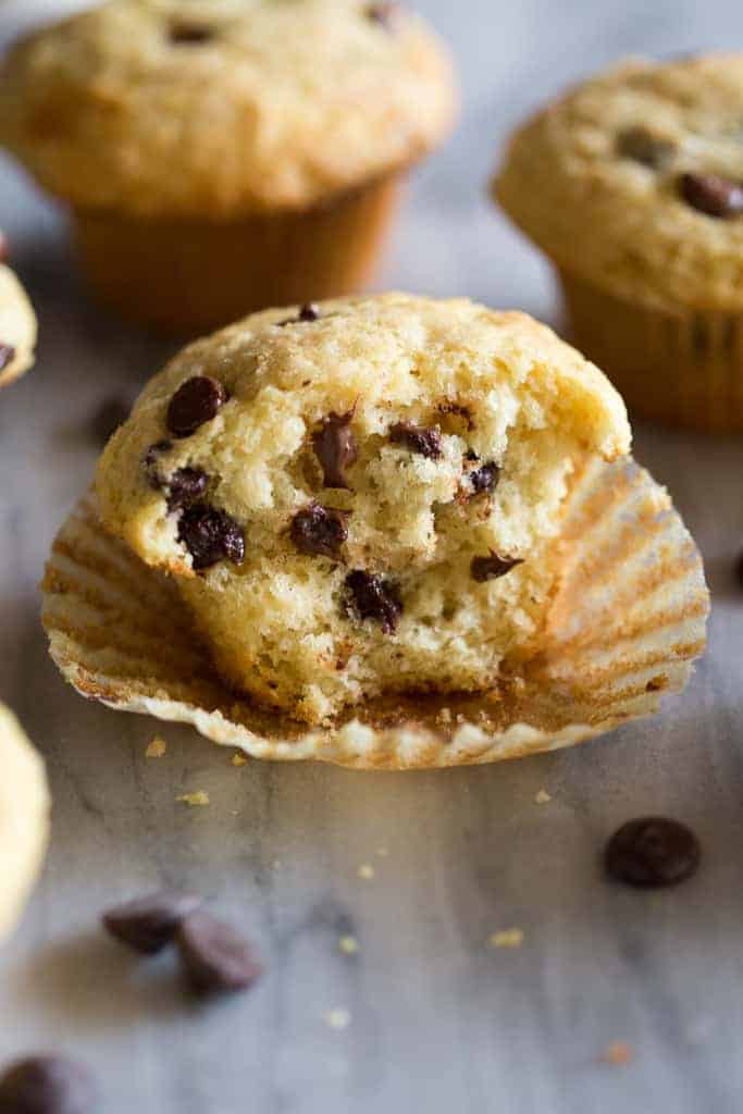 A half eaten chocolate chip muffin with the paper liner pulled down and more muffins in the background.