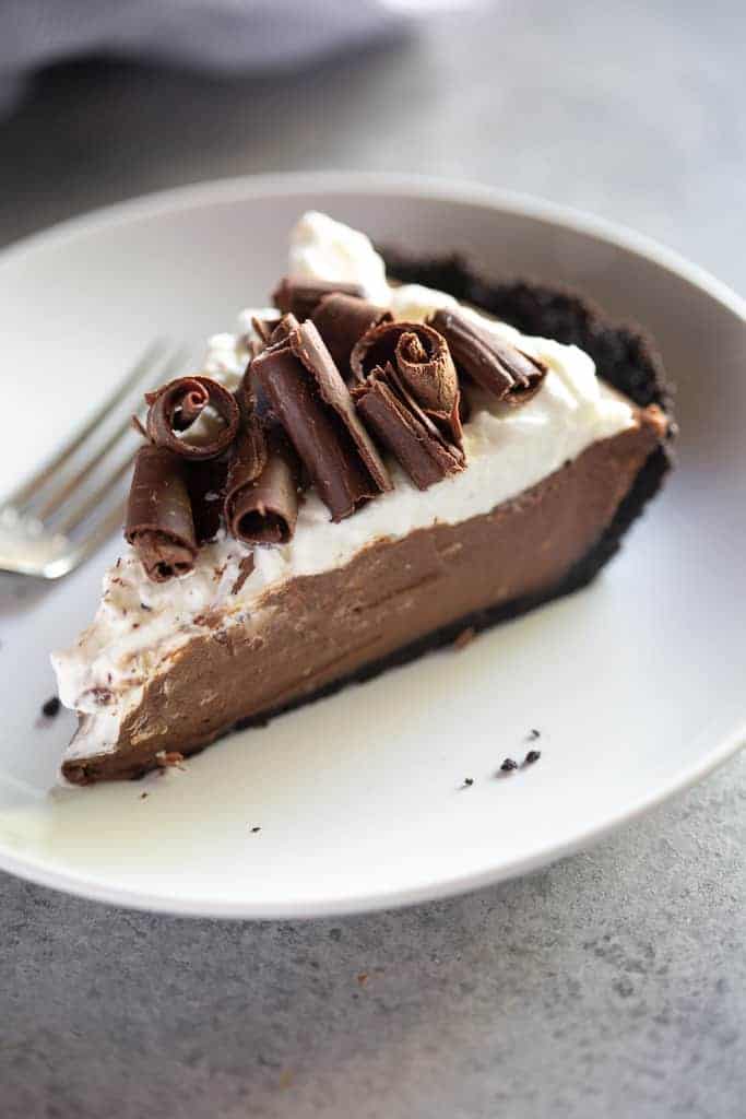 A slice of chocolate cream pie with whipped cream and chocolate curls on top, on a white plate with a fork.