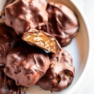 A plate of homemade Chocolate Turtles, one of them cut in half to show the caramel pecan filling.