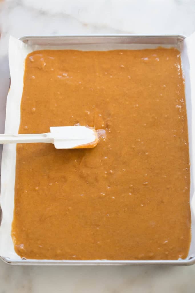 A jelly roll pan lined with parchment paper and pumpkin roll filling being spread over top.