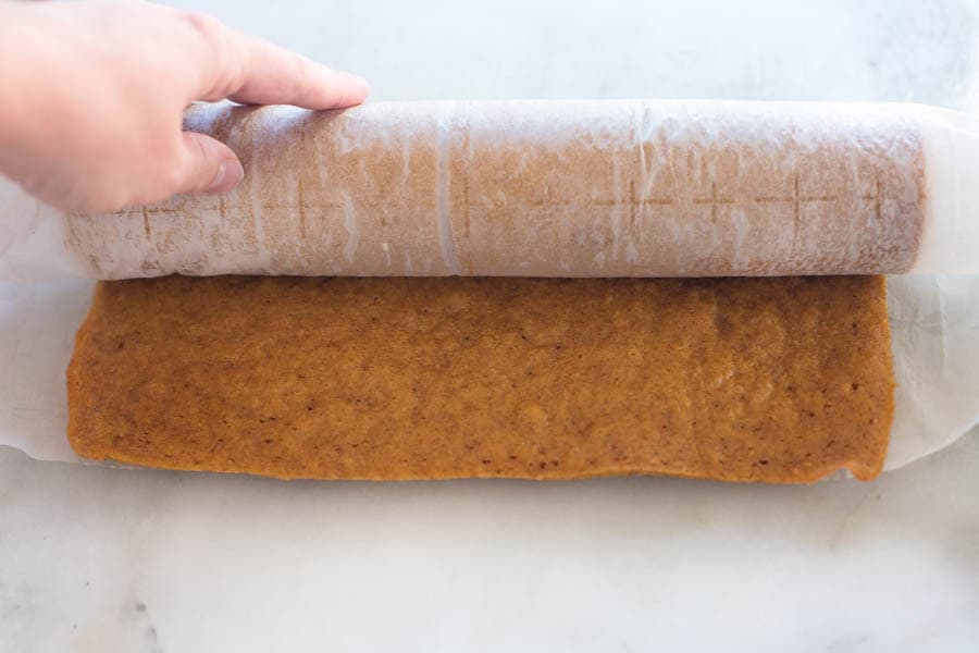 A cooked pumpkin roll, laying horizontally, and being rolled in parchment paper.