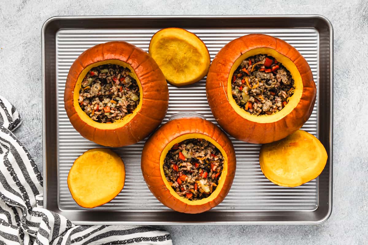 Three pie pumpkins on a baking sheet filled with a mixture of ground sausage, wild rice, and vegetables, ready to be baked.