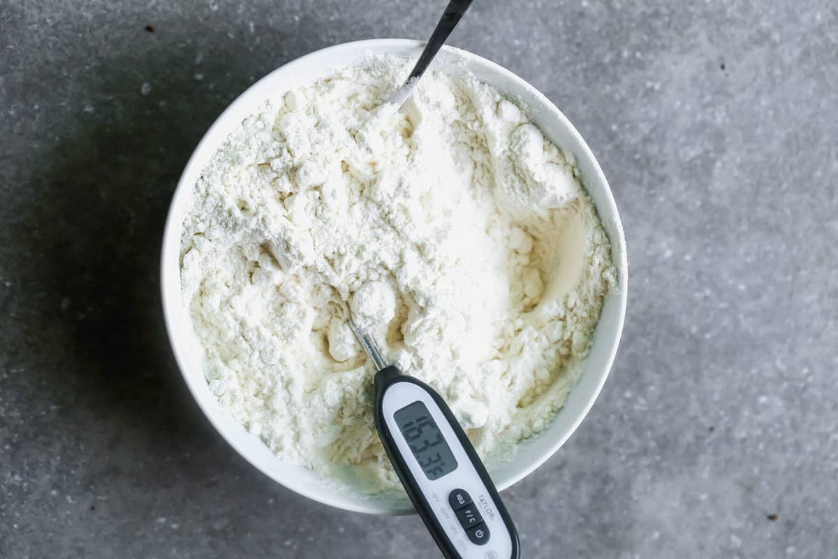 A white mixing bowl filled with flour with a thermometer sticking out to show it has been heat treated.