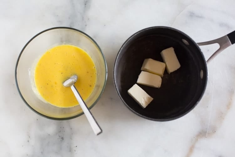 Overhead photo of a clear glass bowl with beaten eggs yolks, cream, lime juice, salt and pepper, next to a saucepan with four tablespoons of butter in it.