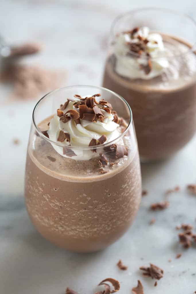 A glass filled with thick frozen hot chocolate, topped with whipped cream and shaved chocolate curls, with a second filled cup in the background.