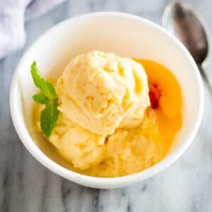 Frozen yogurt served in a white bowl with a sprig of mint and fresh peach slices on the side.