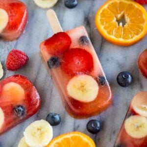 Homemade popsicles made with blueberries, strawberries, bananas and juice, laying on a marble board with fresh fruit scattered around them.