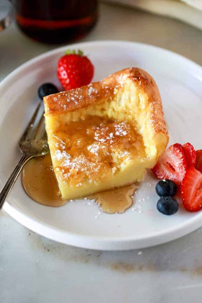 A slice of German Pancakes on a white plate with powdered sugar and syrup on top, served with strawberries and blueberries.