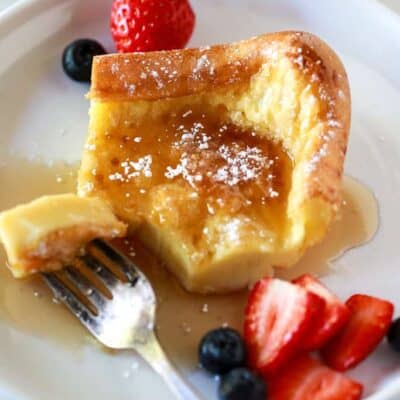 A German pancake on a plate with a fork taking a bite out, served with syrup, powdered sugar and berries.