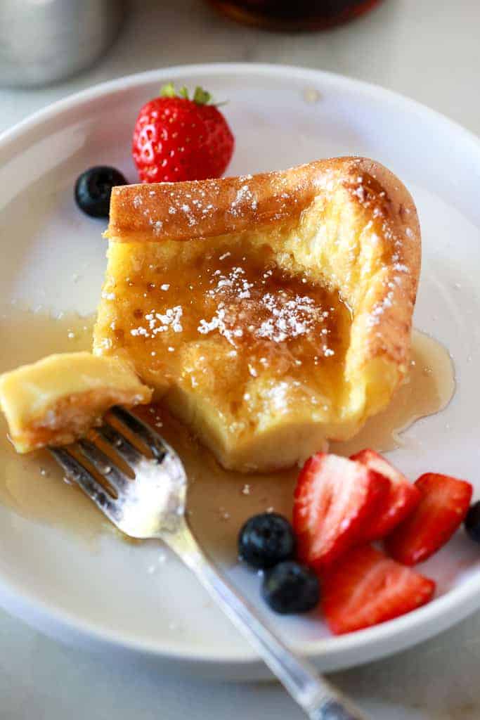 A German pancake on a plate with a fork taking a bite out, served with syrup, powdered sugar and berries.