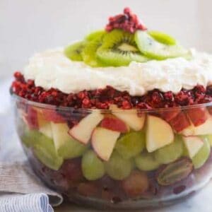 Side profile of a clear bowl filled with fruit including red and green grapes, apples, cranberries, whipped cream and kiwi.