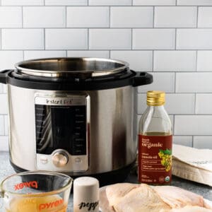 A picture showing an Instant Pot on a counter with canola oil, chicken broth, salt and pepper, and some raw chicken thighs.