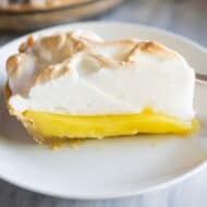 A slice of Lemon Meringue Pie served on a white plate with the remaining pie in the background.