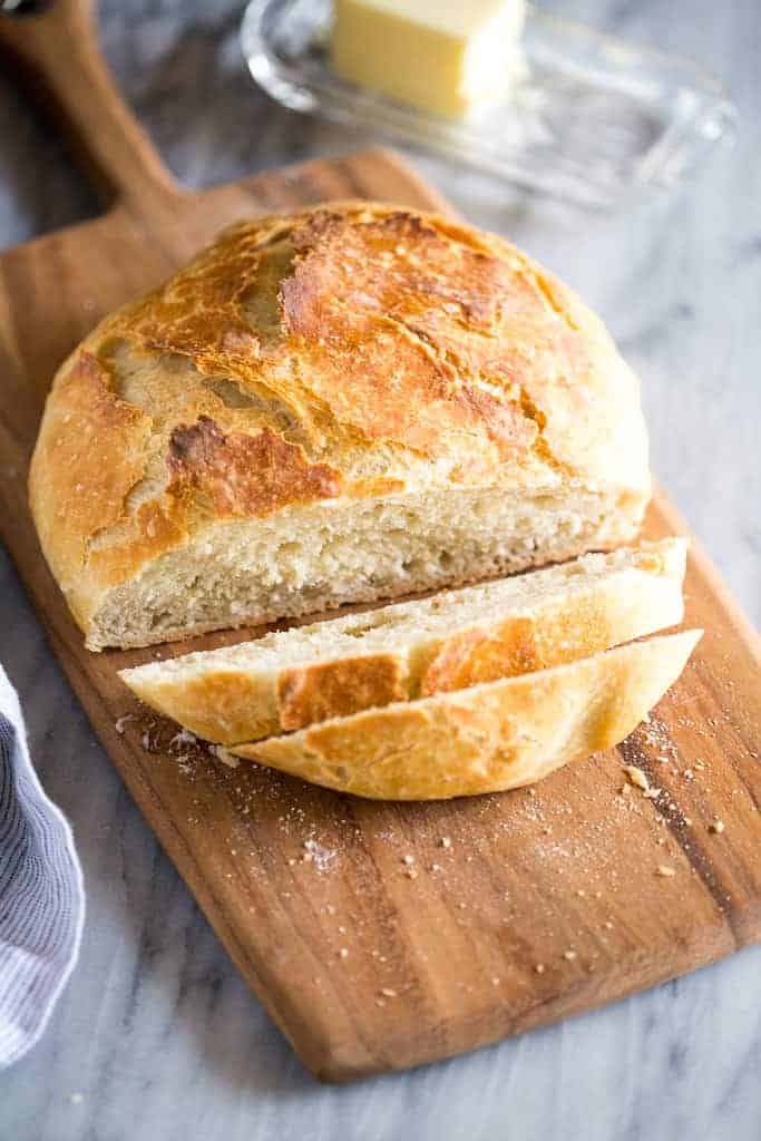 No Knead Bread on a wood cutting board with two slices cut from it and a butter tray in the background.