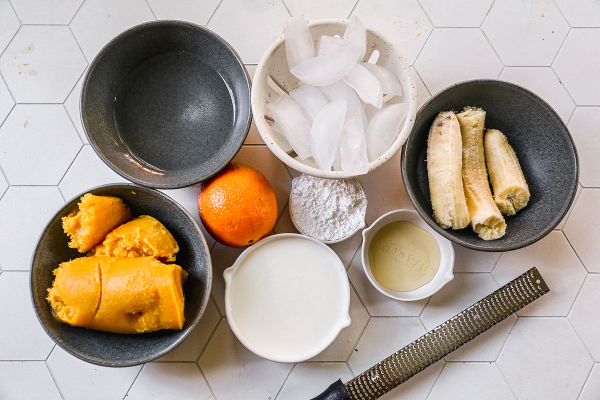 All of the ingredients needed for the best Orange Julius: orange juice concentrate, water, milk, powdered sugar, vanilla, orange zest, banana, and ice.