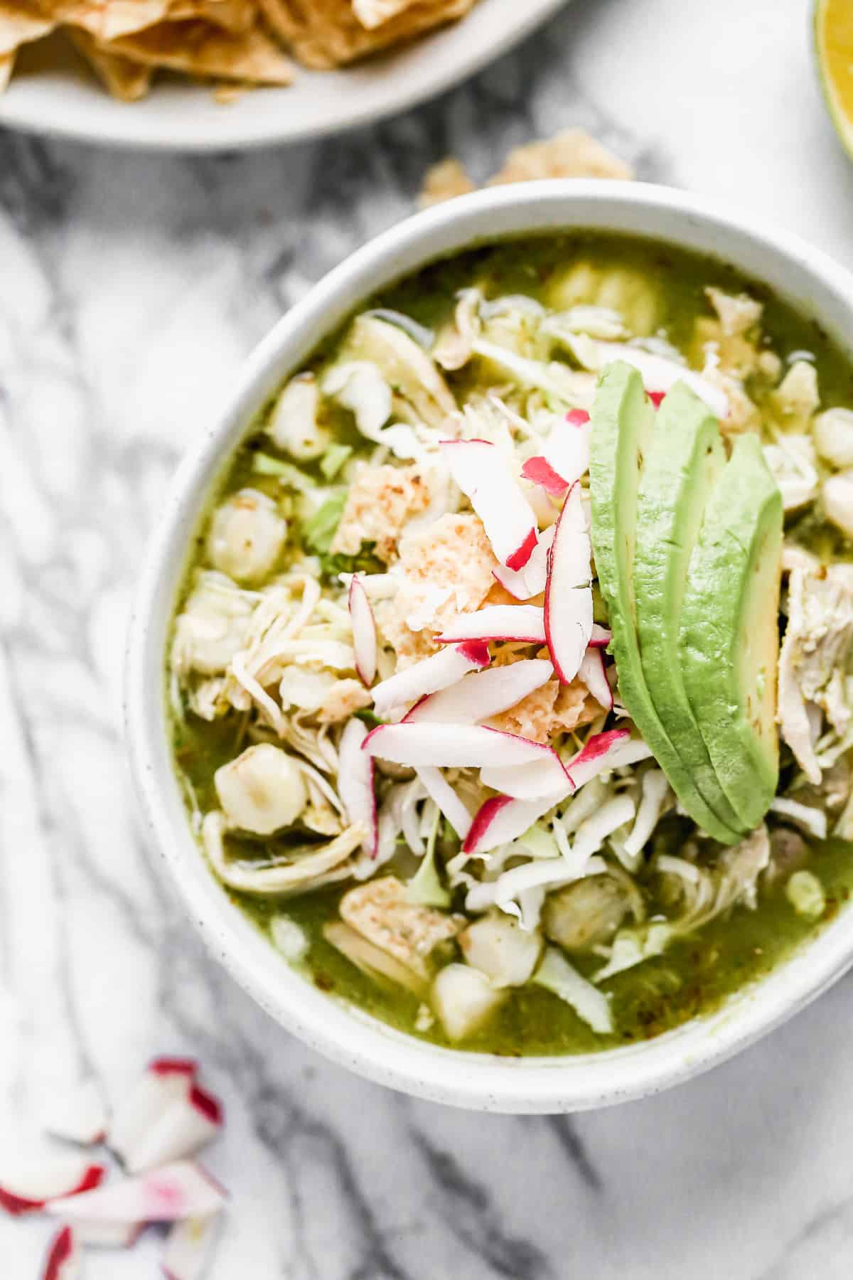 A bowl of Chicken Pozole Verde topped with sliced radishes, crushed tortilla chips, and avocado.