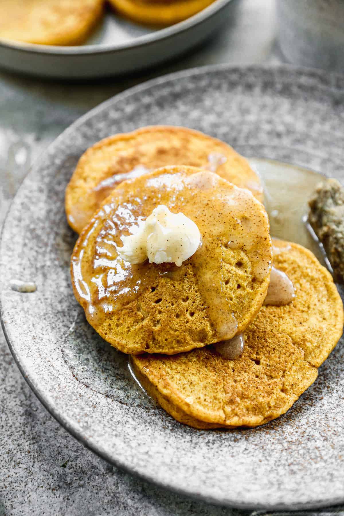 Three homemade Pumpkin Pancakes stacked on top of each other with a spoonful of butter on top and syrup drizzled.