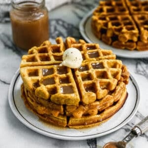 A stack of three Pumpkin Waffles on a plate topped with butter and homemade cinnamon syrup.