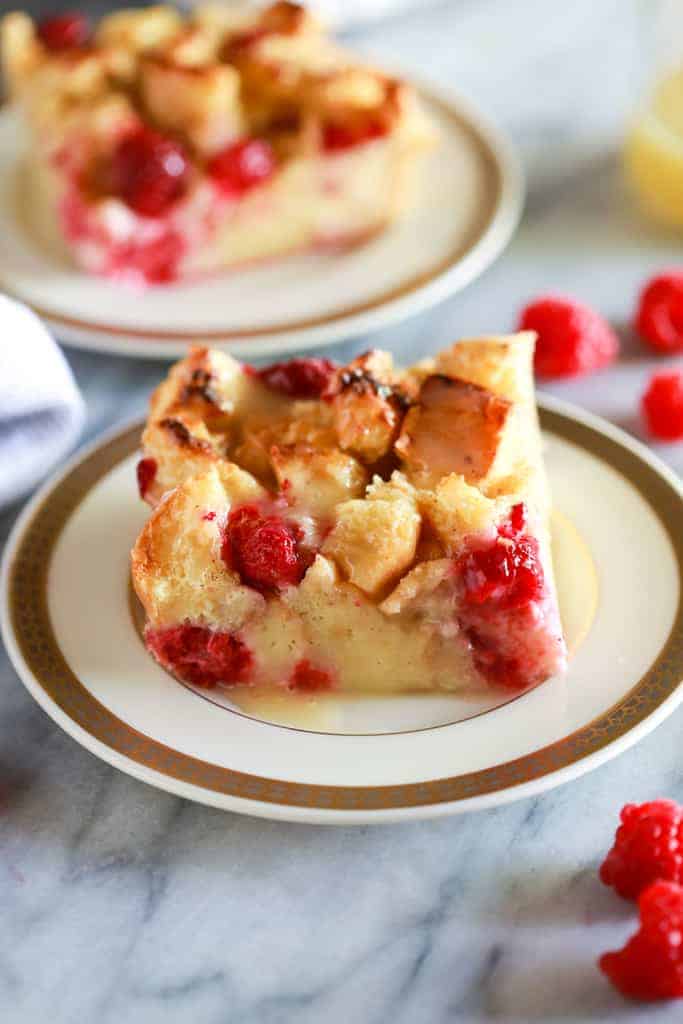 Classic Bread Pudding with raspberries in it and a vanilla cream sauce on top, served on white plate with gold trim and another serving plate in the background.