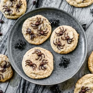 Three chocolate chip Spider Cookies on a plate with toy spiders.