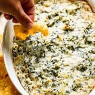 Spinach Artichoke Dip baked in pan, and a hand dipping a chip into it.