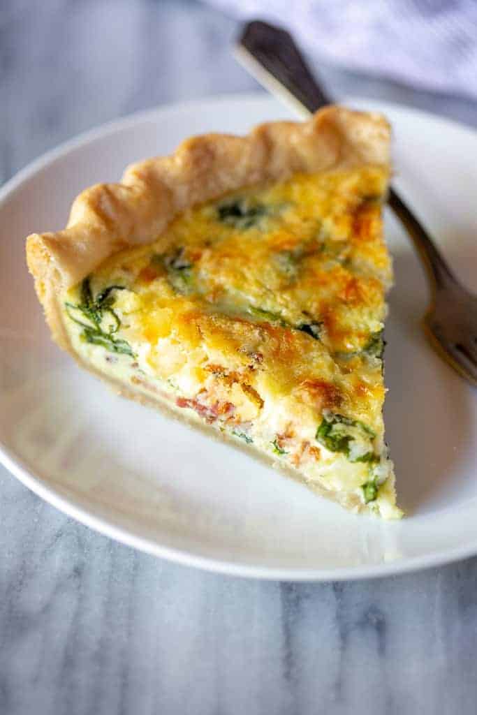 A slice of spinach and bacon quiche on a plate with a fork.