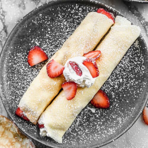 Two homemade Strawberry Crepes topped with whipped cream, strawberries, and a dusting of powdered sugar.