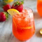 A glass of strawberry lemonade with a strawberry and lemon slice on the cup rim and strawberries and lemons in the background.