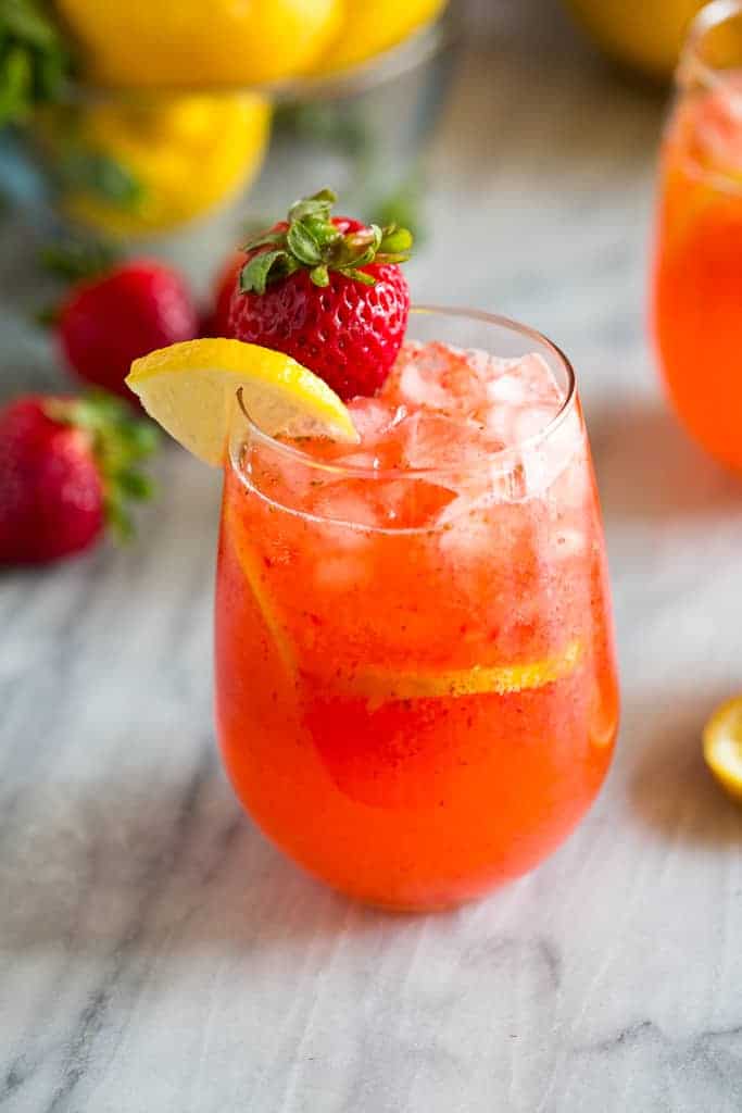 A glass of strawberry lemonade with a strawberry and lemon slice on the cup rim and strawberries and lemons in the background.