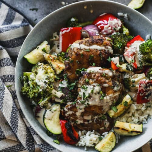 A homemade Chicken Tahini Bowl with grilled chicken and veggies on a bed of rice, and drizzled with tahini sauce and herbs.