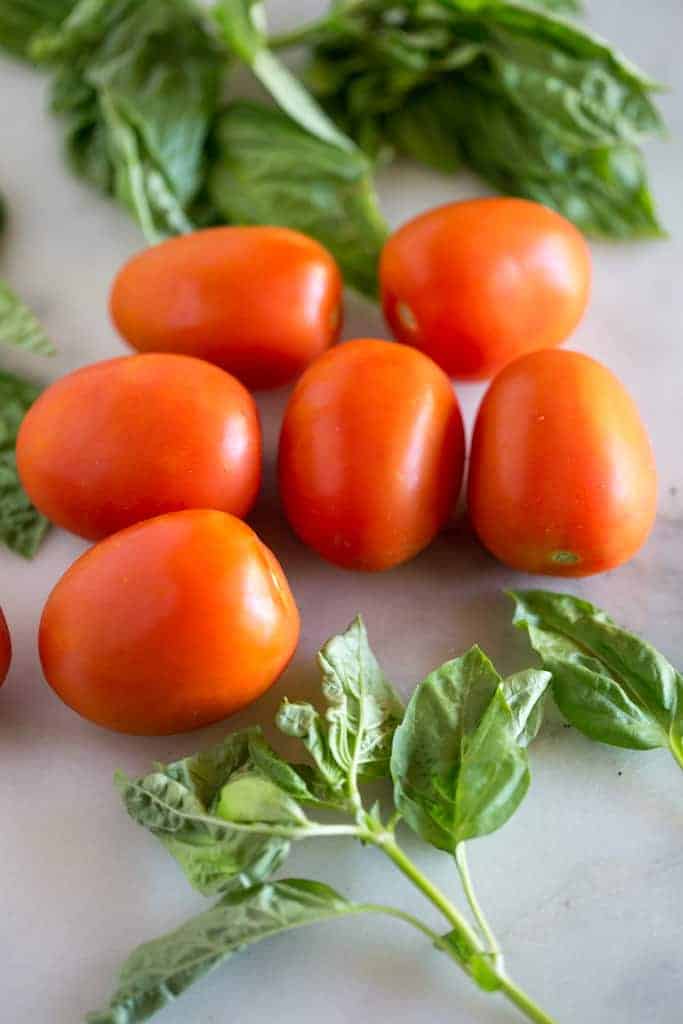 Roma tomatoes on a white marble board with fresh basil leaves.