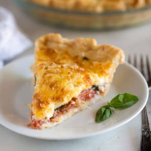 A slice of tomato pie on a white plate with a fork next to it and the rest of the tomato pie in the background.