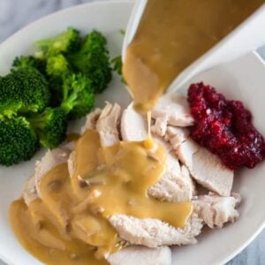 Turkey gravy in a white gravy boat being poured over turkey slices on a plate with cranberry sauce and broccoli.